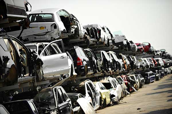 Cars stacked high in a salvage yard, many with visible damage, under a bright sky.