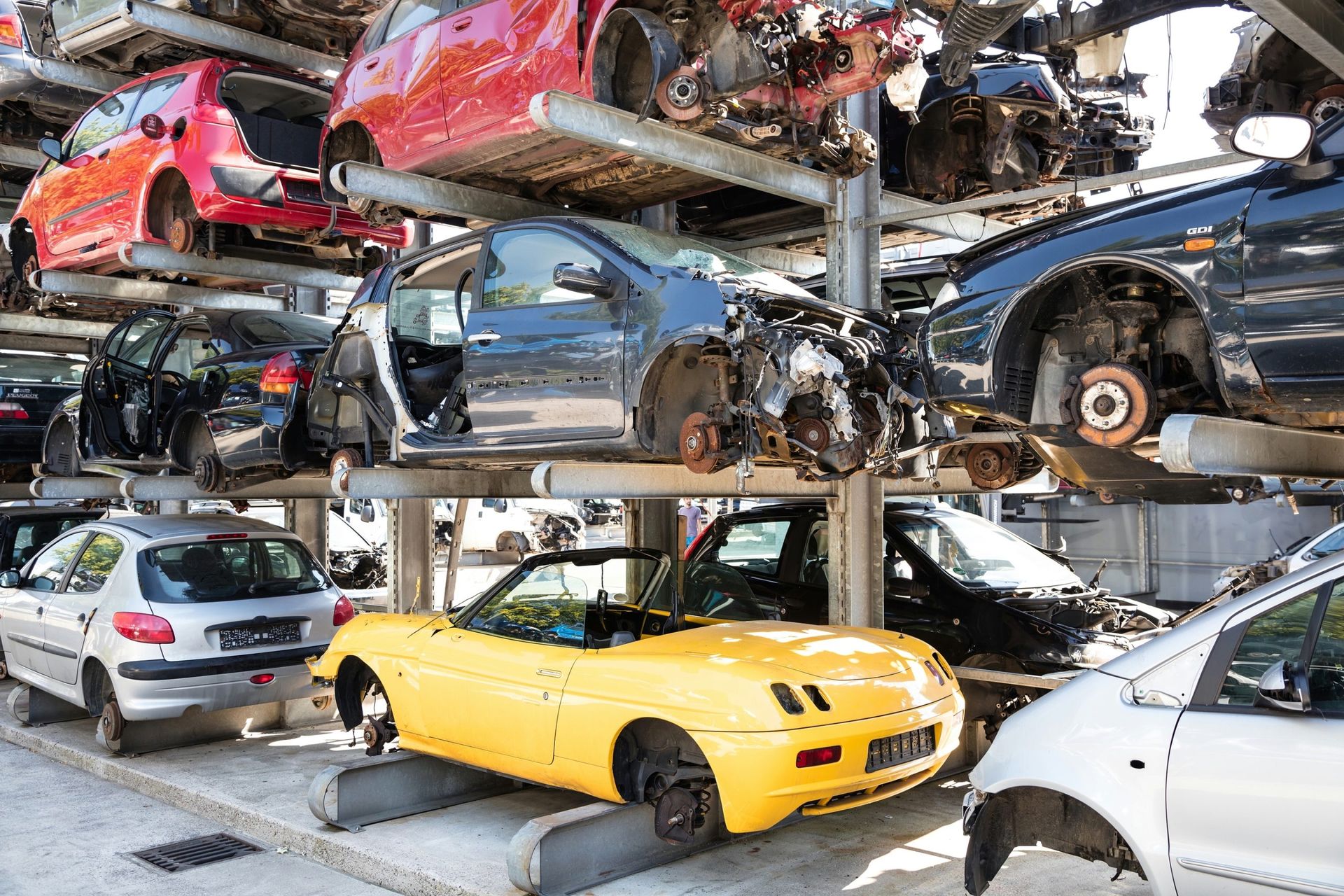 Cars stacked on a multi-level rack in a salvage yard; mostly damaged, various colors.