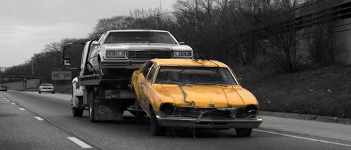 Tow truck carrying a yellow car and a white car on a highway. The scene is mostly black and white, with the cars in color.
