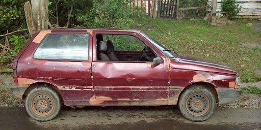 A dilapidated, maroon Fiat Uno parked on the side of a road, next to a green grassy hill.